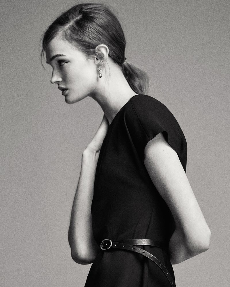 Profile view of a young woman with fair skin and a black dress in a neutral-toned studio setting. Fashion shot with a model.