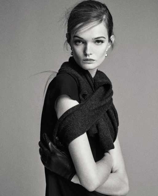 Black and white portrait of a fashionable woman wearing a black dress, leather gloves, and a dark shawl against a neutral background.