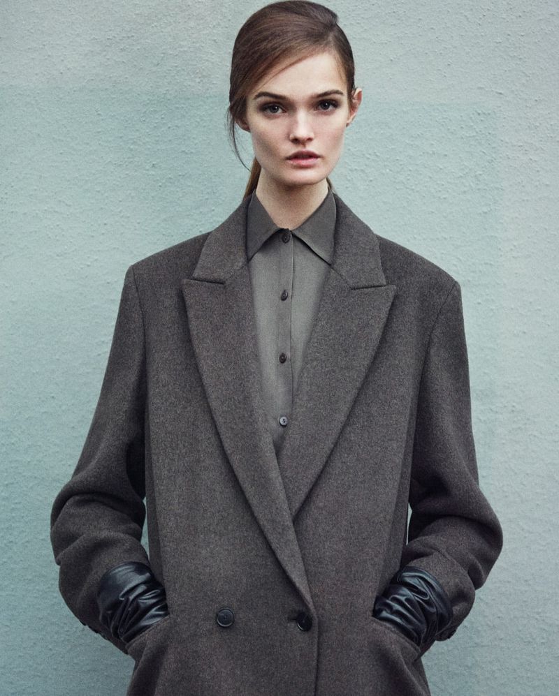 Fashion shot of a model in a grey coat and leather gloves looking at the camera against a plain blue wall in a studio setting.