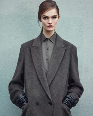 Fashion shot of a model in a grey coat and leather gloves looking at the camera against a plain blue wall in a studio setting.