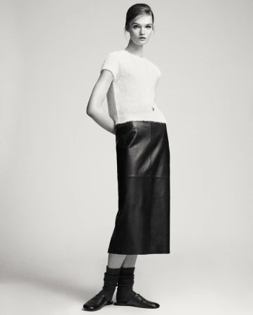 Model Lulu Tenney wearing a white textured top and black leather skirt against a seamless light grey studio background.