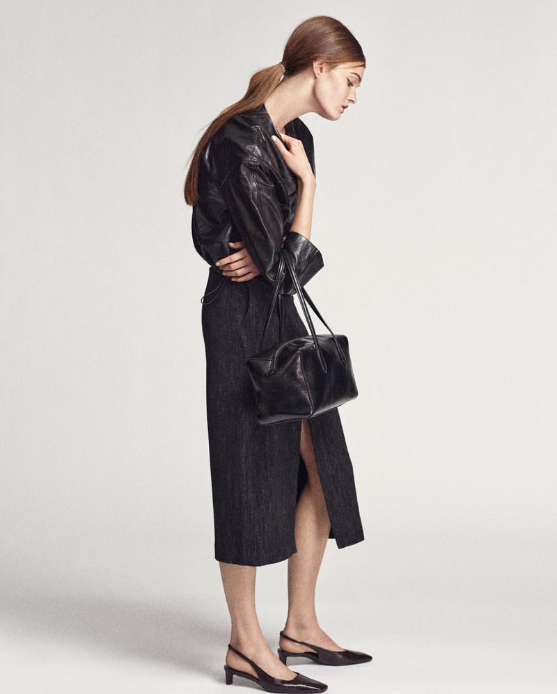 Woman wearing a black jacket, dark midi skirt, and black slingback heels carrying a black purse, posing in a light studio.