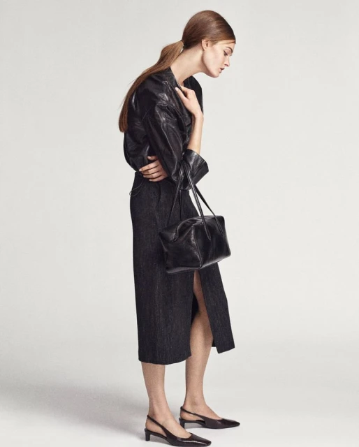 Woman wearing a black jacket, dark midi skirt, and black slingback heels carrying a black purse, posing in a light studio.