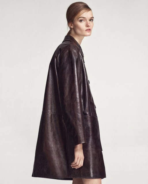 A woman in a brown leather coat poses indoors. The shot features neutral lighting and emphasizes the coat's design and texture.