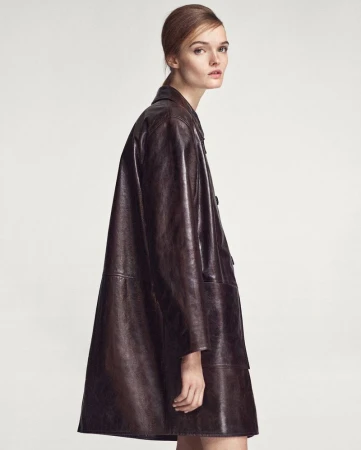 A woman in a brown leather coat poses indoors. The shot features neutral lighting and emphasizes the coat's design and texture.