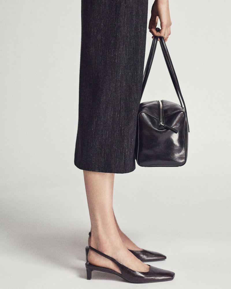 A woman models a square, black leather purse with short handles, paired with a gray midi skirt and black slingback heels against a plain background.