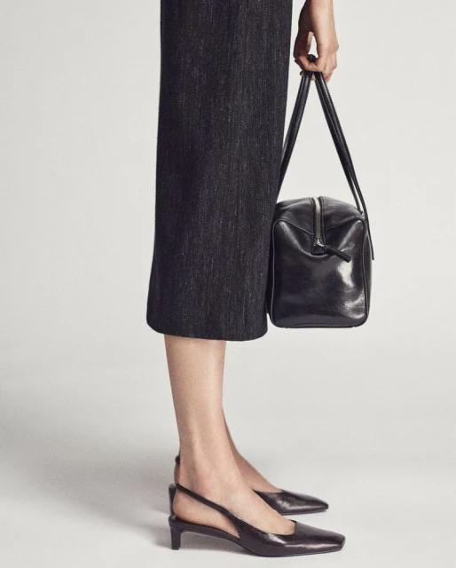 A woman models a square, black leather purse with short handles, paired with a gray midi skirt and black slingback heels against a plain background.