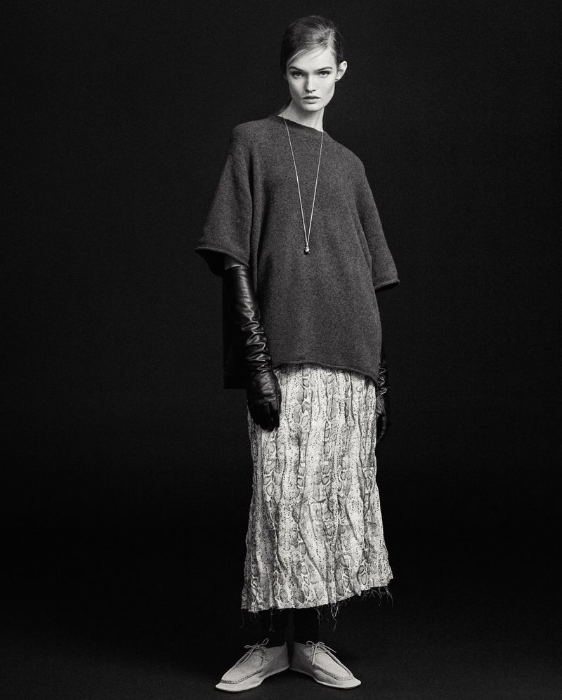 Black and white studio shot of a stylish female fashion model wearing a gray sweater, patterned skirt, long gloves, and neutral shoes.