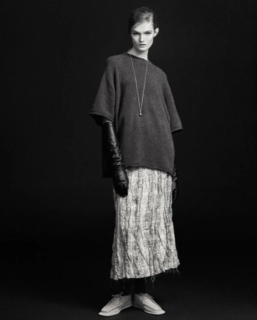Black and white studio shot of a stylish female fashion model wearing a gray sweater, patterned skirt, long gloves, and neutral shoes.