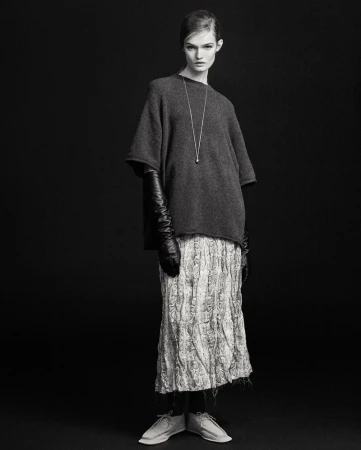 Black and white studio shot of a stylish female fashion model wearing a gray sweater, patterned skirt, long gloves, and neutral shoes.