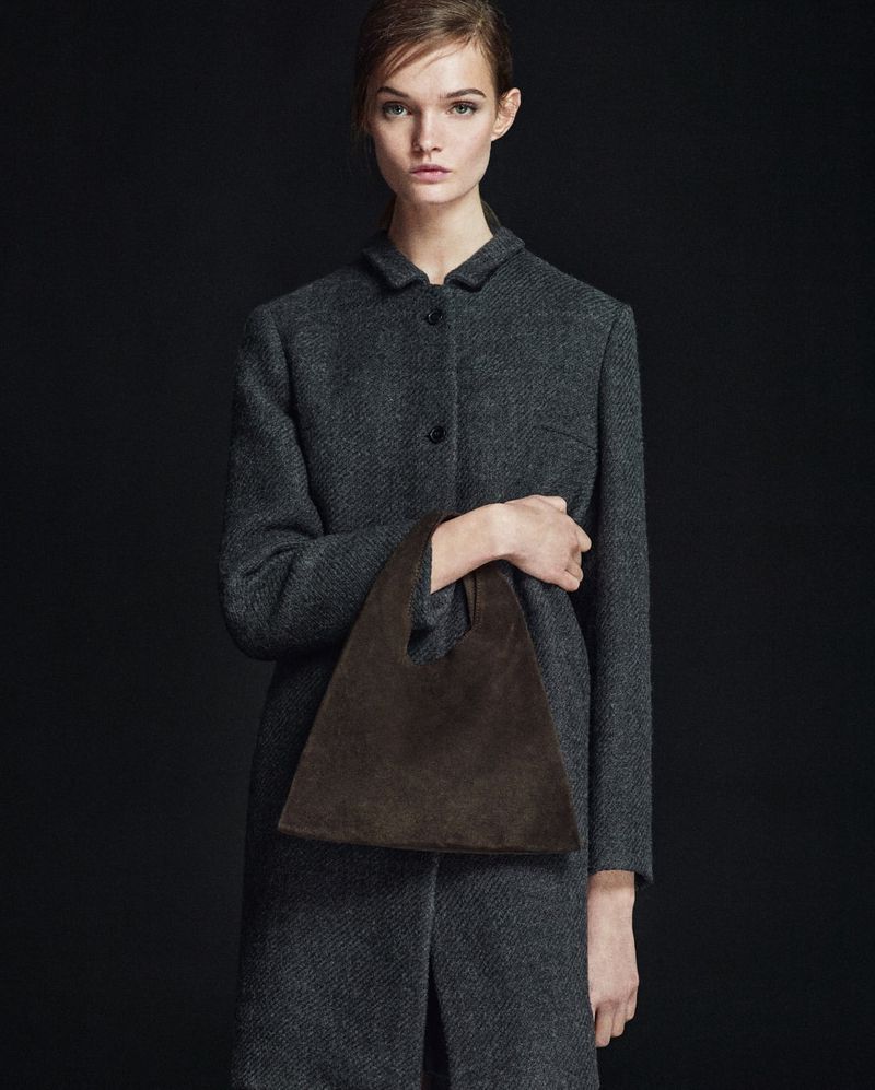 Fashion shot of woman in grey coat holding a brown suede handbag against dark background. Elegant, minimalist style. Ideal for fashion blogs.