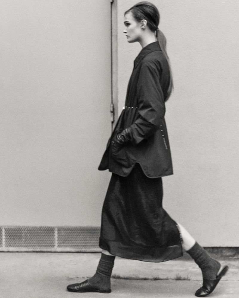 Monochrome fashion shot of a woman in coat and dress walking against a light wall, showcasing editorial style and minimal color palette.
