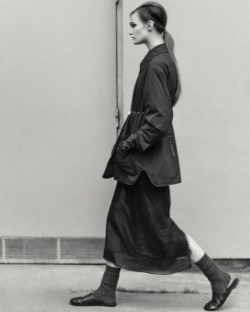 Monochrome fashion shot of a woman in coat and dress walking against a light wall, showcasing editorial style and minimal color palette.