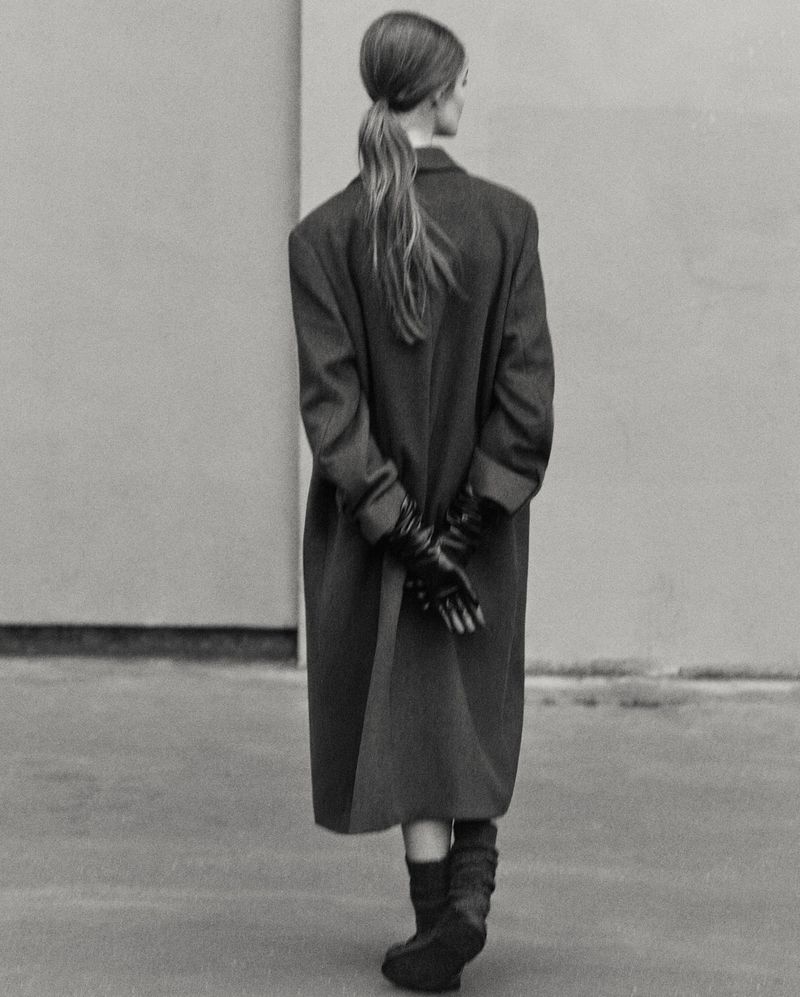 Model Lulu Tenney from behind wearing an oversized dark coat, gloves, and boots against a plain wall for Massimo Dutti.