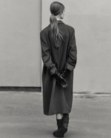 Model Lulu Tenney from behind wearing an oversized dark coat, gloves, and boots against a plain wall for Massimo Dutti.