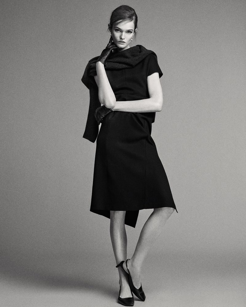 A full shot captures a woman posing in a black dress, gloves and shoes against a grey backdrop in a fashion photography studio setting.