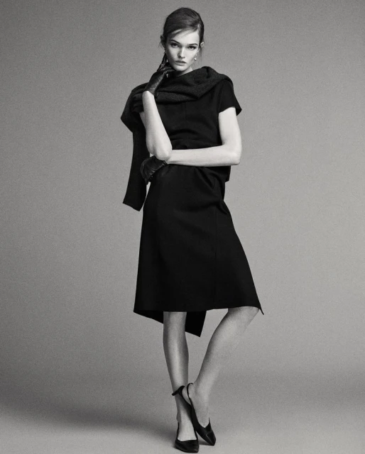 A full shot captures a woman posing in a black dress, gloves and shoes against a grey backdrop in a fashion photography studio setting.