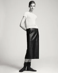 Model Lulu Tenney wearing a white textured top and black leather skirt against a seamless light grey studio background.