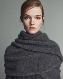 Close-up portrait of female model Lulu Tenney wearing a dark grey textured wool scarf against a solid grey studio backdrop.