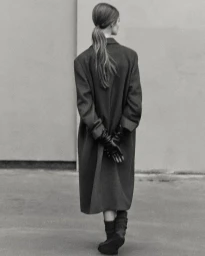 Model Lulu Tenney from behind wearing an oversized dark coat, gloves, and boots against a plain wall for Massimo Dutti.