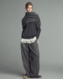 Full-length portrait of a model wearing a dark grey sweater with a draped scarf and wide-leg trousers against a grey backdrop.