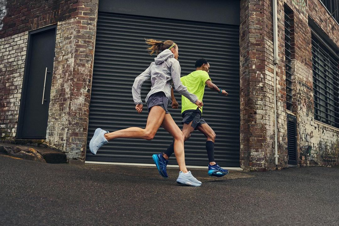 Two athletes running on an asphalt road alongside a brick building with a metal door during daylight, with focus on motion and athleticism.