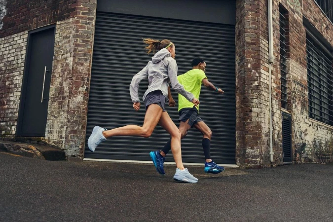 Two athletes running on an asphalt road alongside a brick building with a metal door during daylight, with focus on motion and athleticism.