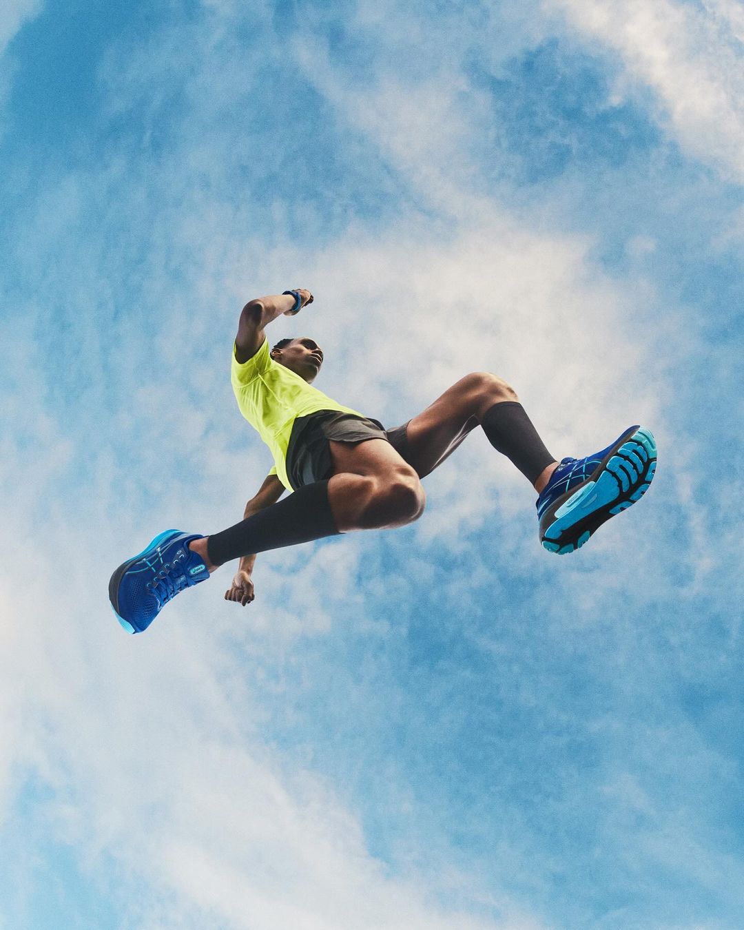 Low angle view of athletic male in sportswear jumping against a blue sky with clouds, showcasing fitness and dynamic movement.