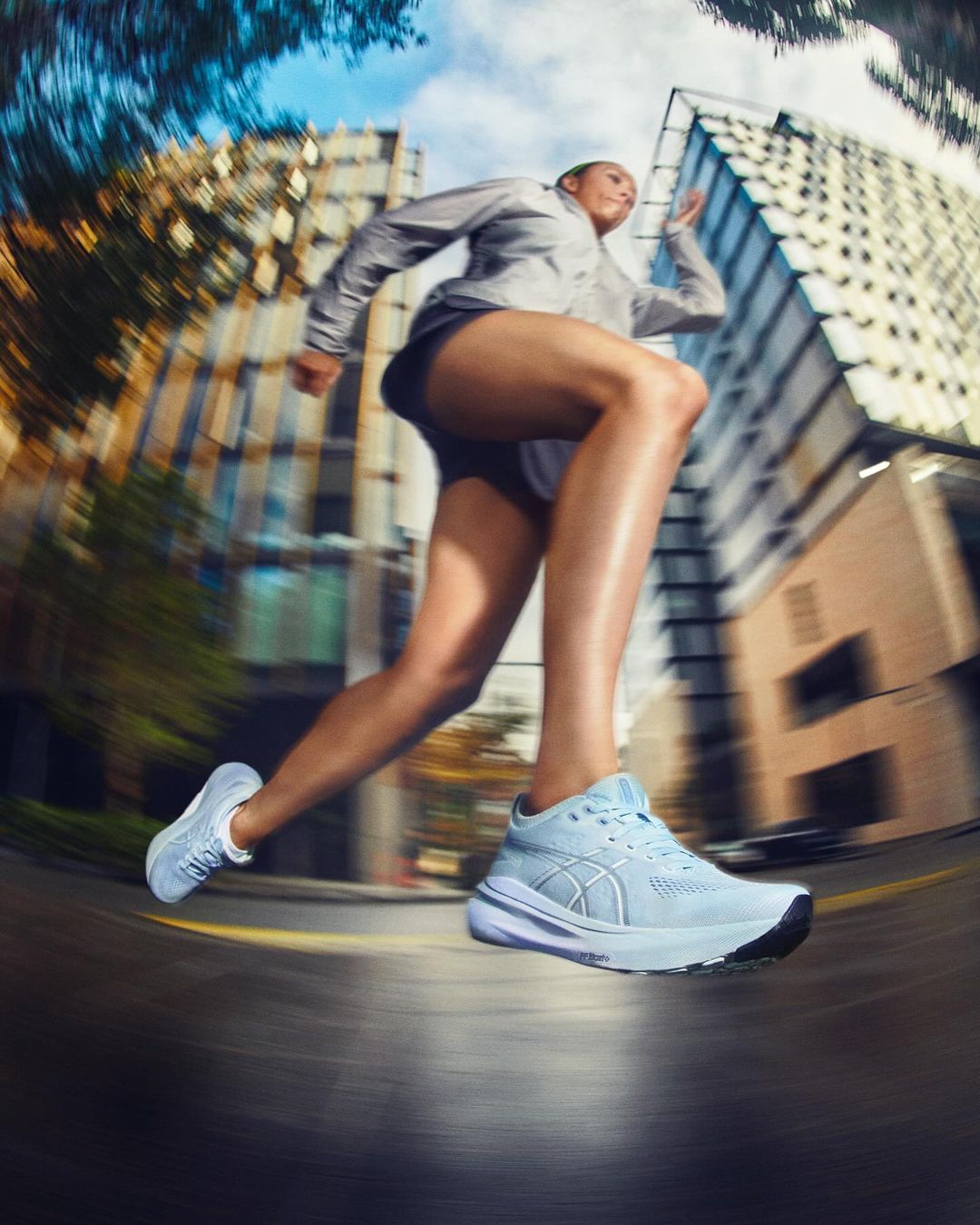Woman running on a city street wearing grey athletic wear and light blue running shoes with blurred motion and tall buildings in the background.