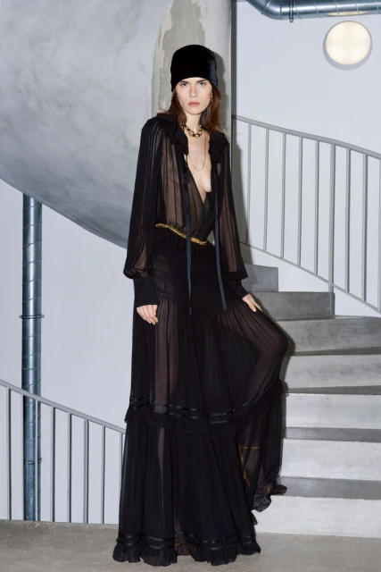 A woman in a long, sheer black dress and a black headscarf stands on concrete stairs with metal railings in an indoor setting.