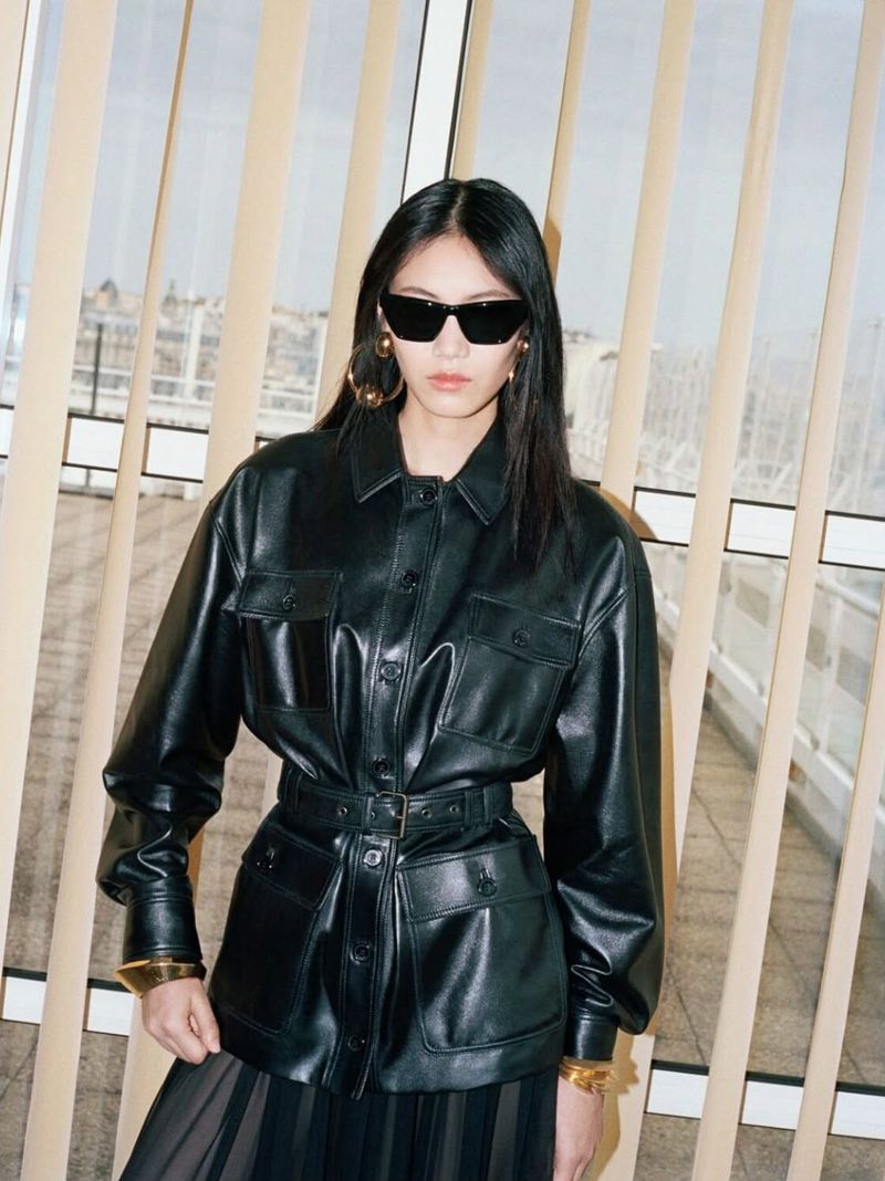 An Asian woman poses in a belted black leather jacket and sunglasses, showcasing a fashion-forward look against a neutral background.