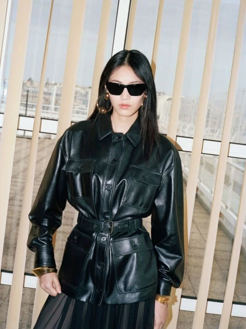An Asian woman poses in a belted black leather jacket and sunglasses, showcasing a fashion-forward look against a neutral background.