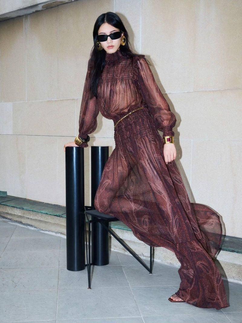 Model wearing dark sunglasses and a sheer brown patterned dress posing near a chair against a stone wall.