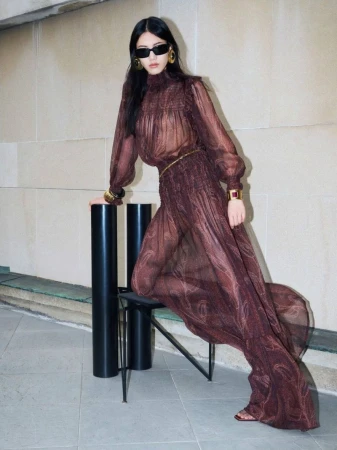 Model wearing dark sunglasses and a sheer brown patterned dress posing near a chair against a stone wall.