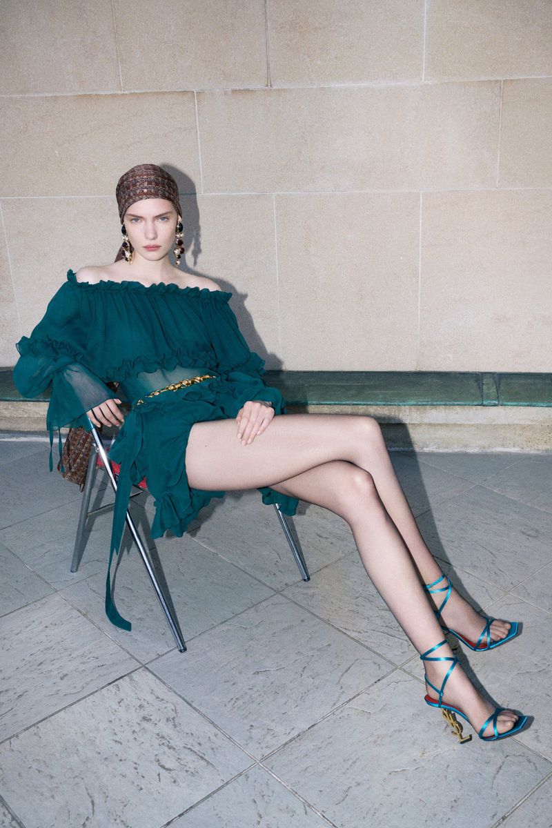 Fashionable woman in a teal dress with a patterned headscarf sitting on a chair against a neutral tiled wall, showcasing an elegant pose.