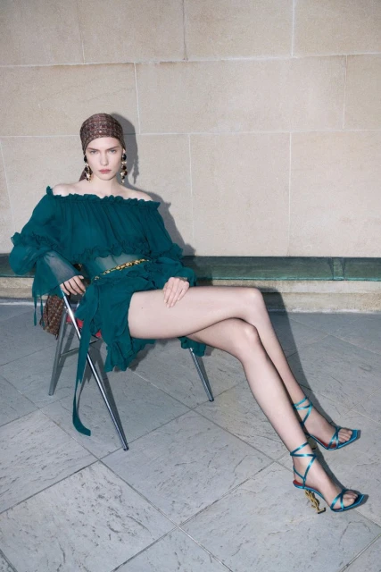 Fashionable woman in a teal dress with a patterned headscarf sitting on a chair against a neutral tiled wall, showcasing an elegant pose.
