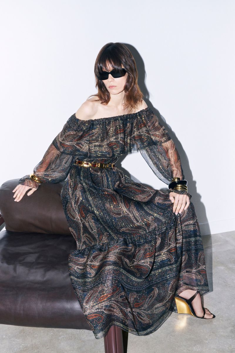 A brunette woman in an off-the-shoulder paisley maxi dress with sunglasses, gold accessories, and black heeled sandals, sits indoors.