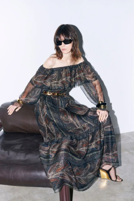 A brunette woman in an off-the-shoulder paisley maxi dress with sunglasses, gold accessories, and black heeled sandals, sits indoors.