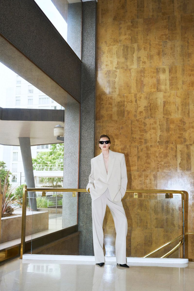 Fashionable adult woman in a cream-colored suit and sunglasses stands on a polished floor near brass rails and a decorative wall.