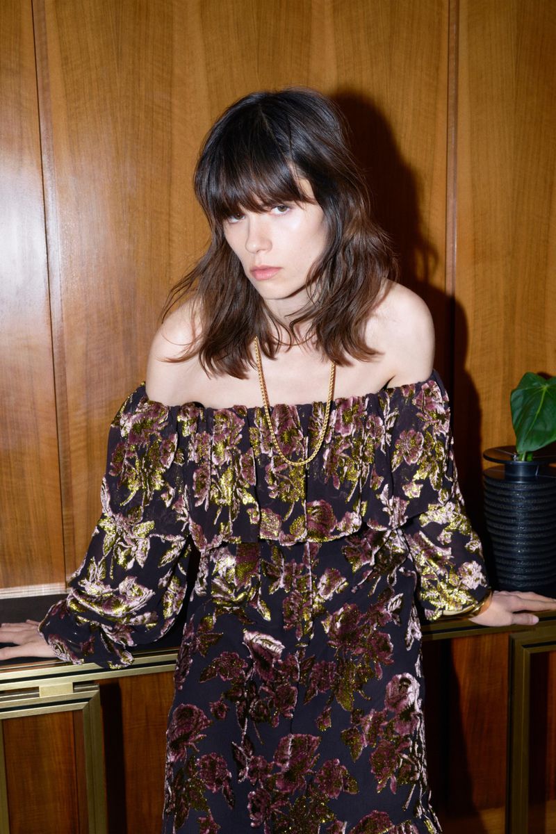 Adult woman in an off-the-shoulder floral dress poses indoors against a wood paneled wall with a dark plant pot on a console table.