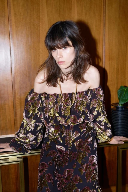 Adult woman in an off-the-shoulder floral dress poses indoors against a wood paneled wall with a dark plant pot on a console table.