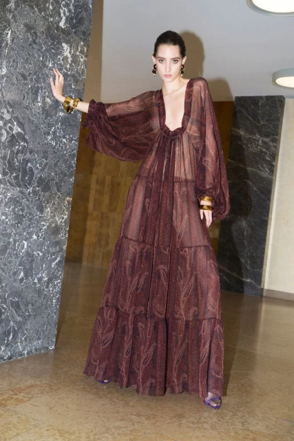 A woman in a long brown gown with full sleeves, wearing gold bracelets, poses indoors near a marble wall, casting a shadow.