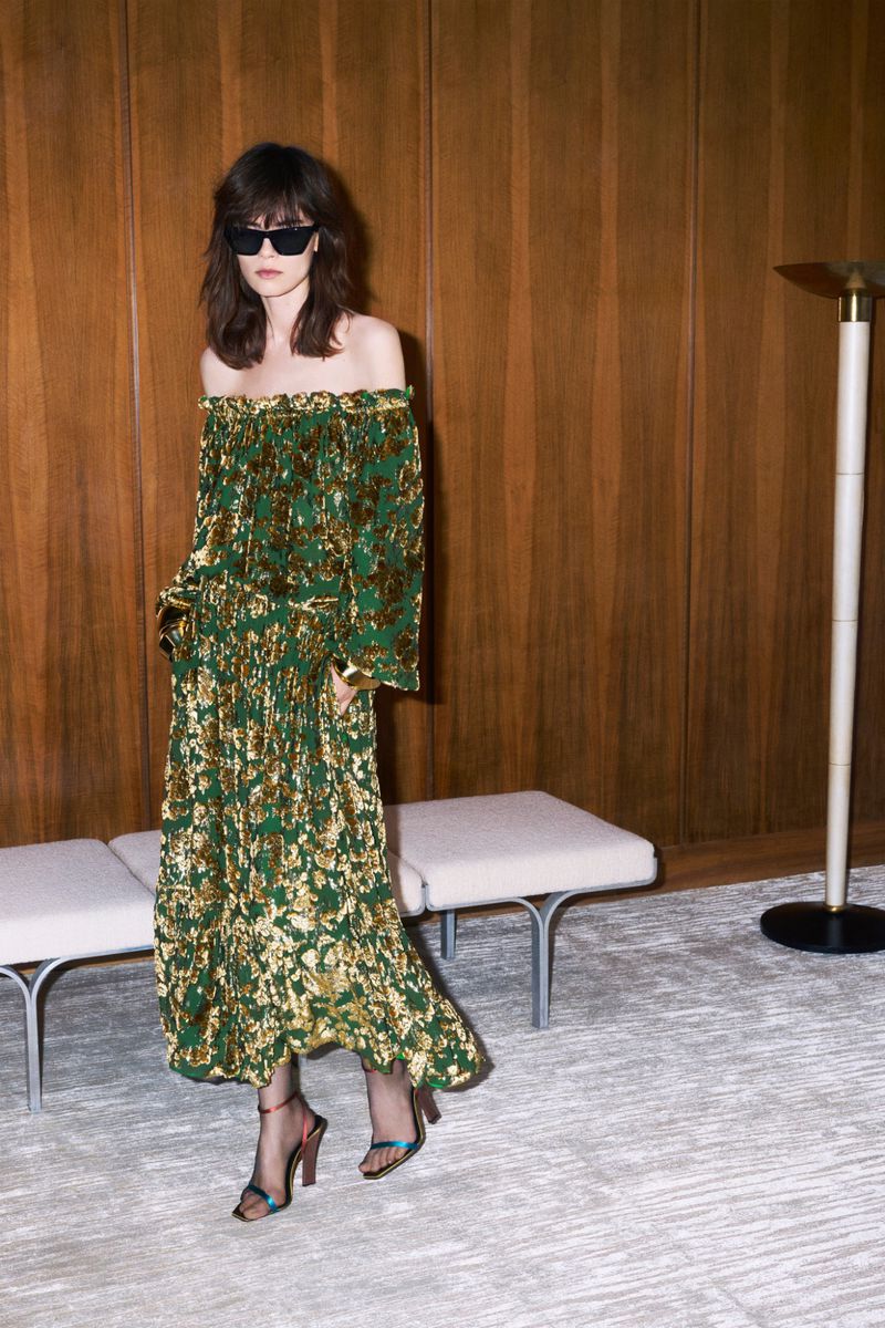 Model in off-the-shoulder patterned dress and sunglasses standing in a wood-paneled room