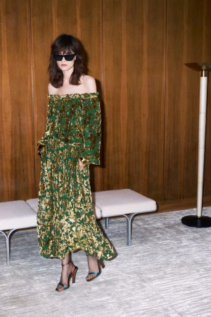 Model in off-the-shoulder patterned dress and sunglasses standing in a wood-paneled room