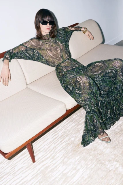 A woman poses on a couch, wearing a flowing green paisley-patterned outfit and black sunglasses, complemented by neutral tones in a modern interior.