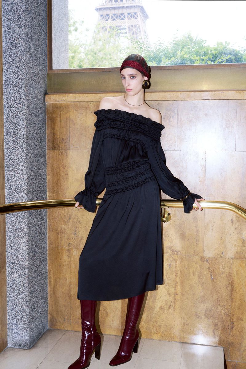 A fashionable woman poses in a black off-the-shoulder dress and high burgundy boots, with the Eiffel Tower visible through a window.