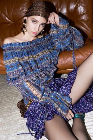 Fashion shot of a young adult woman in a patterned off-the-shoulder top and wrap, seated on a brown leather sofa, wearing statement earrings.