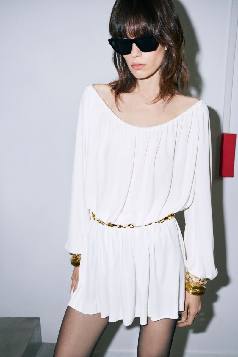 Fashionable woman wearing a long-sleeved white dress, black sunglasses, gold bracelets and a gold chain belt, posing in an indoor setting.