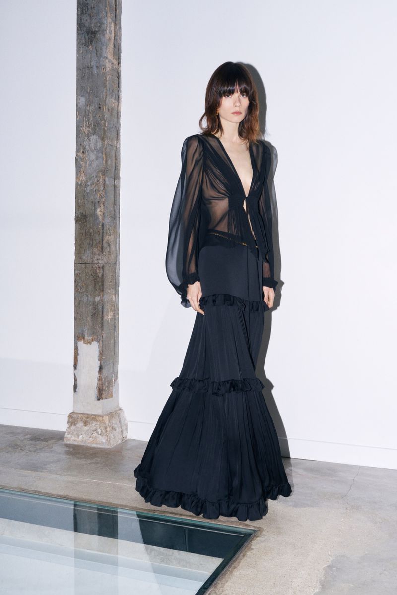 Adult woman in a long black flowing dress with a sheer blouse stands in a minimalist studio with white walls and a concrete floor.