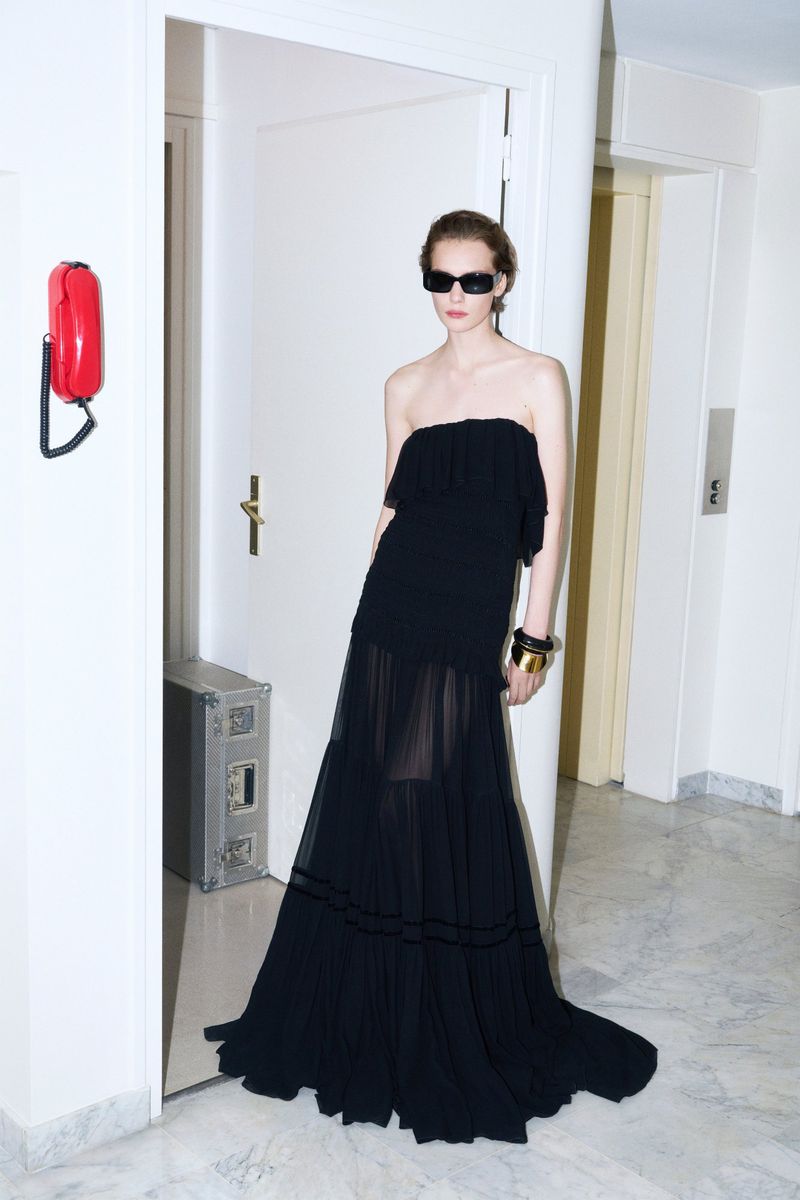 Fashionable adult female posing in a black strapless gown and sunglasses in an indoor setting with a red telephone on the wall.